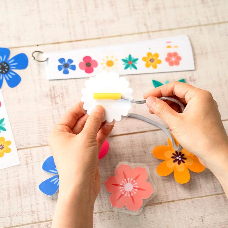 May include: A person is assembling a floral mobile. The mobile features various colorful flower cutouts, including blue, pink, orange, and yellow designs. The mobile is suspended by a gray cord and a metal ring.
