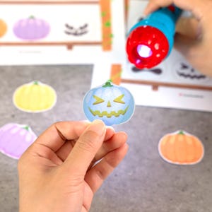 May include: A blue pumpkin-shaped cutout with a yellow jack-o'-lantern face being illuminated by a blue and red flashlight. Additional pumpkin cutouts in yellow, purple, and orange are scattered nearby on a gray surface.