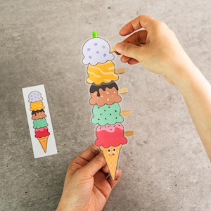 May include: A colorful ice cream cone illustration with six scoops of different flavors, held together by wooden clothespins. The ice cream scoops are in various colors, including pink, green, brown, yellow, and blue. A hand is holding the top clothespin.