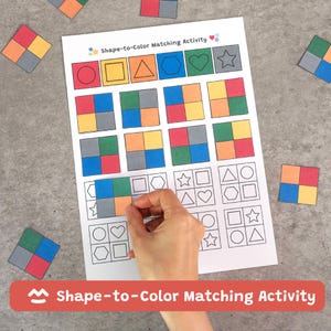 Op de afbeelding: Een Shape-to-Color Matching Activity-spel met een wit vel met vormen en kleuren. Een hand plaatst een vierkant kleurblok. Extra kleurblokken liggen verspreid rond het vel. Het spel is ontworpen voor educatieve doeleinden.
