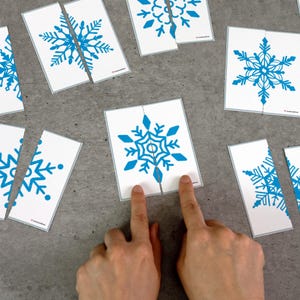 May include: White cards with blue snowflake designs, some cut in half, are scattered on a gray surface. Two fingers are touching a card, attempting to connect the puzzle pieces. The snowflake design is a winter theme.