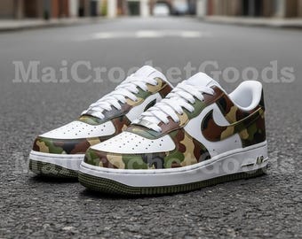 May include: Pair of white and camouflage Air Force 1 sneakers. The sneakers feature a white base with camouflage accents in shades of green, brown, and tan. The Nike swoosh is white. The soles are white with a green trim. The text "MaiCrochetGoods" is visible.