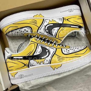 Può includere: Un paio di scarpe da ginnastica gialle e bianche con dettagli neri. Le scarpe presentano un design unico con contorni neri e la scritta "AIR" sul lato. Le scarpe da ginnastica sono in una scatola, il che suggerisce che sono nuove.