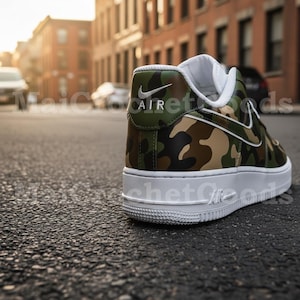 May include: A close-up of a camouflage-patterned sneaker with a white sole. The shoe features a green, brown, and tan camouflage design with a white Nike swoosh. The word "AIR" is visible on the heel.