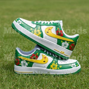 Puede incluir: Un par de zapatillas blancas y verdes con dise&ntilde;os florales y detalles amarillos. Las zapatillas presentan un logotipo amarillo y un dise&ntilde;o de bandera de golf. Los zapatos est&aacute;n colocados sobre una superficie de hierba verde.