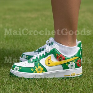 Puede incluir: Zapatillas blancas y verdes con dise&ntilde;os florales y de golf. Las zapatillas presentan una base blanca con detalles verdes, estampados florales amarillos y rojos, y un swoosh amarillo con una bandera de golf. La suela es blanca con un detalle "AR" amarillo.