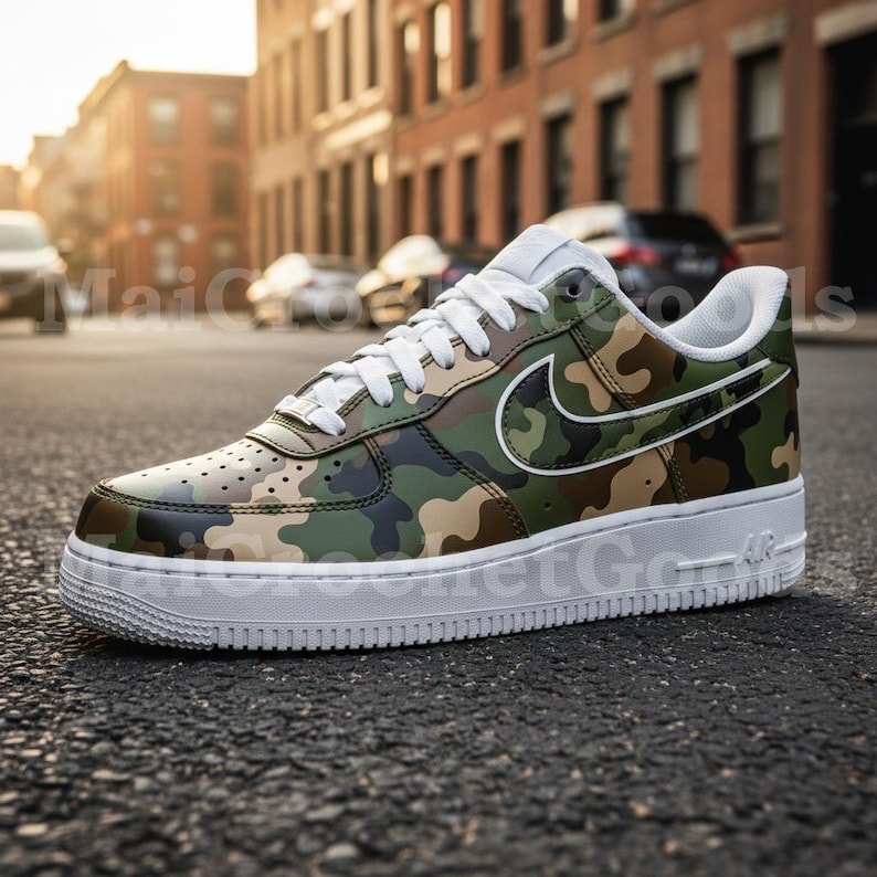 May include: Camouflage-patterned Air Force 1 sneakers with white accents. The shoe features a green, brown, and black camouflage design on the upper, a white Nike swoosh, and a white sole. The laces are white, and the shoe is set on an asphalt surface.