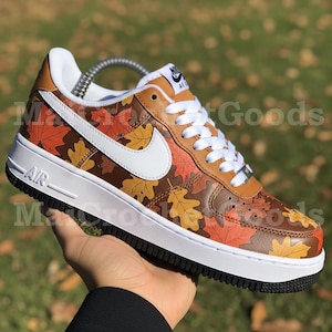Puede incluir: Zapatillas Nike Air Force 1 con diseño de hojas de otoño. Las zapatillas tienen suela blanca, cordones blancos y un logo Nike blanco. La parte superior está decorada con hojas marrones, naranjas y amarillas. La palabra "AIR" está impresa en el lateral.