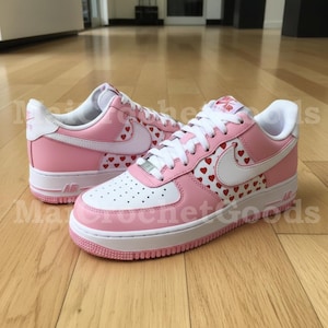 Könnte beinhalten: Paar rosa-weiße Sneaker mit roten Herzmustern. Die Schuhe haben weiße Schnürsenkel, einen weißen Nike-Swoosh und eine weiße Sohle. Die Sneaker stehen auf einem Holzboden.