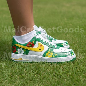Puede incluir: Zapatillas blancas y verdes con dise&ntilde;os florales y de golf. Las zapatillas presentan un logotipo amarillo con una bandera de golf, flores de hibisco rojas y blancas y la palabra "AIR" en dorado.