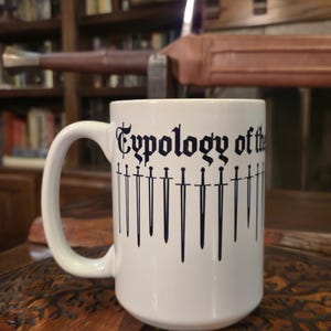 Può includere: Tazza in ceramica bianca con la scritta "Typology of the" in carattere gotico, con illustrazioni di spade nere. La tazza ha un manico grande ed è appoggiata su una superficie di legno.