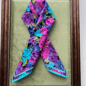 May include: A framed vintage scarf with a floral pattern. The scarf features a dark background with vibrant purple, pink, and turquoise flowers. The scarf is framed in a dark brown wooden frame with a green background.