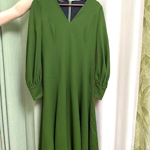 Pode incluir: Vestido verde-oliva de mangas compridas com decote em V. O vestido tem uma saia rodada e está pendurado num cabide de madeira. As mangas são franzidas nos ombros e terminam em punhos justos. O vestido tem fecho de correr.