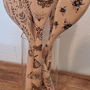 May include: A set of wooden kitchen utensils with engraved insect and plant designs. The utensils, including spoons and spatulas, are displayed in a clear glass container, resting on a wooden surface. The designs include butterflies, bees, and foliage.