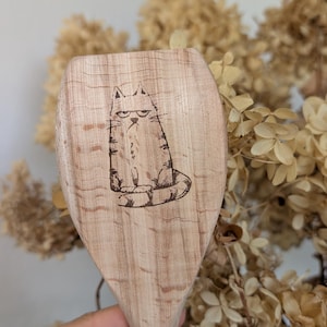 Op de afbeelding: Houten lepel met een lasergeëtste illustratie van een chagrijnige kat. De lepel is gemaakt van lichtgekleurd hout en heeft een brede, afgeronde kop. De kat wordt zittend afgebeeld met een strenge uitdrukking. De lepel wordt in een hand gehouden.