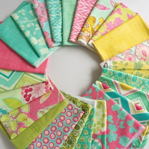 May include: A circular display of folded fabric squares in a variety of colours and patterns. The fabrics feature shades of pink, green, yellow, and turquoise, with floral, geometric, and abstract designs. The arrangement is on a white surface.