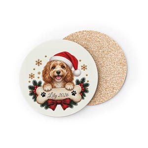 May include: A round coaster with a cork base and a white top featuring a brown dog wearing a red Santa hat. The dog rests its paws on a bone-shaped graphic labelled Lily 2026, surrounded by pine branches, red berries, snowflakes, and a red plaid bow.