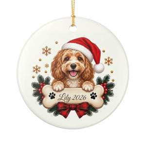 May include: A round white ceramic ornament featuring a brown curly-haired dog wearing a red Santa hat. The dog rests its paws on a bone-shaped sign with the text Lily 2026, flanked by pine branches, red berries, a red plaid bow, and gold snowflakes.