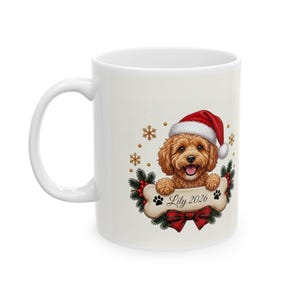 May include: A white ceramic mug featuring a graphic of a brown curly-haired dog wearing a red and white Santa hat. The dog rests its paws on a bone-shaped sign with the text Lily 2026, surrounded by pine branches, red berries, and gold snowflakes.