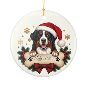 May include: A round white ceramic ornament featuring a Bernese Mountain Dog wearing a red Santa hat. The dog rests its paws on a bone-shaped sign inscribed with the name Lily and the year 2026, flanked by pine branches, red berries, and a plaid bow.