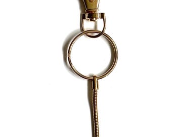 Pode incluir: Um porta-chaves dourado com um mosquetão, um anel grande e uma extensão flexível em espiral que termina numa pequena bola. Projetado para prender chaves ou outros itens pequenos.