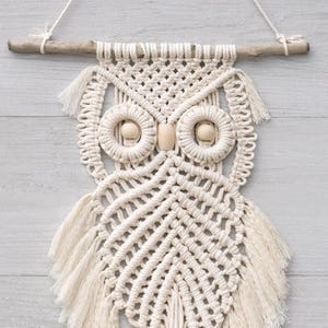 May include: Cream-colored macrame owl wall hanging. The owl features a detailed woven design with wooden bead eyes and a wooden dowel base. The owl is suspended from a natural wooden branch and a knotted cord.