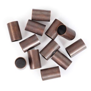 May include: A group of 12 copper-colored metal tubes, all the same size and shape, are scattered on a white surface. The tubes are cylindrical with rounded edges.