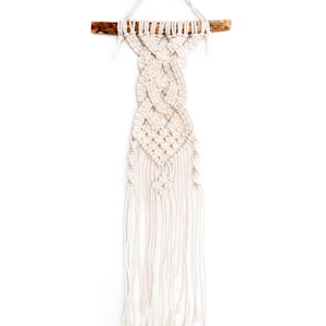 May include: A white macrame wall hanging with a wooden dowel at the top. The macrame is made with a knot pattern and has long fringe at the bottom.