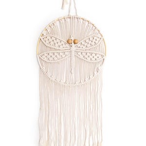 May include: A white macrame wall hanging with a dragonfly design and wooden beads. The hanging has a circular wooden hoop and long white fringe.