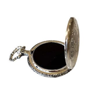 May include: Silver pocket watch with an open lid, revealing a black interior. The watch has a decorative design on the lid and a detailed handle. The watch is set against a white background.