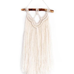 Puede incluir: Un tapiz de macramé blanco con un patrón de diamante y flecos. El tapiz está sujeto a una rama de madera natural.