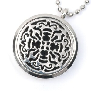 Puede incluir: Un collar de plata con un colgante redondo de diseño calado. El colgante presenta un patrón negro intrincado con un motivo floral. El collar está hecho de una cadena de plata.