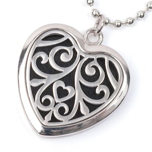 Puede incluir: Collar con colgante de corazón plateado con un diseño de filigrana negro. El colgante tiene un diseño recortado que revela el fondo negro.