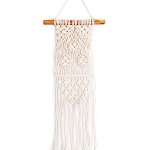 Puede incluir: Un tapiz de macramé blanco con una barra de madera en la parte superior. El macramé tiene un patrón geométrico y una larga franja.