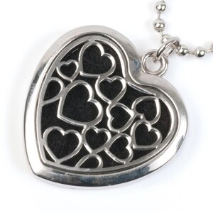 Puede incluir: Un collar con un colgante en forma de corazón de plata con un diseño delicado de corazones más pequeños recortados. El colgante tiene un fondo negro.