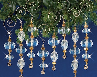 Op de afbeelding: Acht transparante glazen kerstversieringen met gouden kralen en draadhangers. De versieringen hangen aan een groene dennentak tegen een blauwe achtergrond.