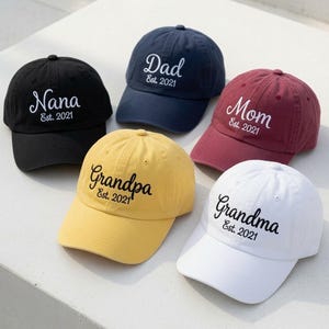 May include: A collection of five baseball caps in black, navy blue, maroon, yellow, and white. Each cap features embroidered text including Nana, Dad, Mom, Grandpa, or Grandma, followed by the text Est. 2021 in a cursive and sans-serif font style.