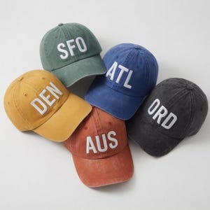 May include: A collection of six baseball caps in various colors, including green, blue, yellow, red, and gray. Each cap features a different three-letter city abbreviation embroidered in white on the front.