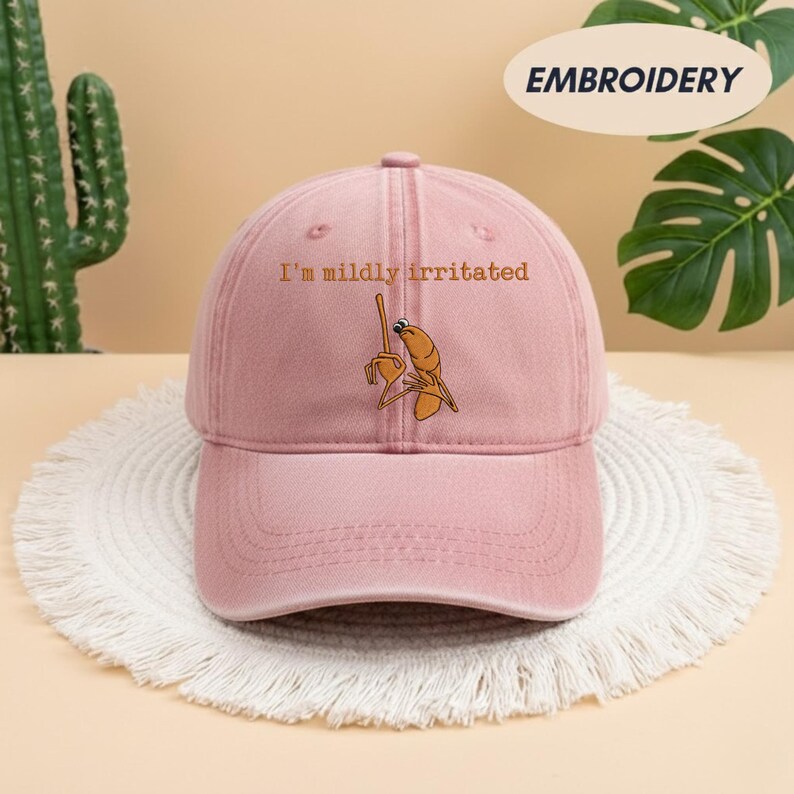 May include: A dusty rose baseball cap with the embroidered phrase "I'm mildly irritated" above a cartoon shrimp character. The cap has a curved brim and a fabric strap closure.