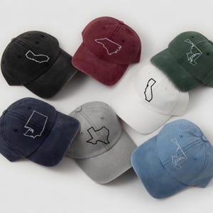 May include: A collection of baseball caps in various colors, each embroidered with a state outline. Colors include black, burgundy, navy, gray, white, green, and blue. The caps are arranged on a white surface.
