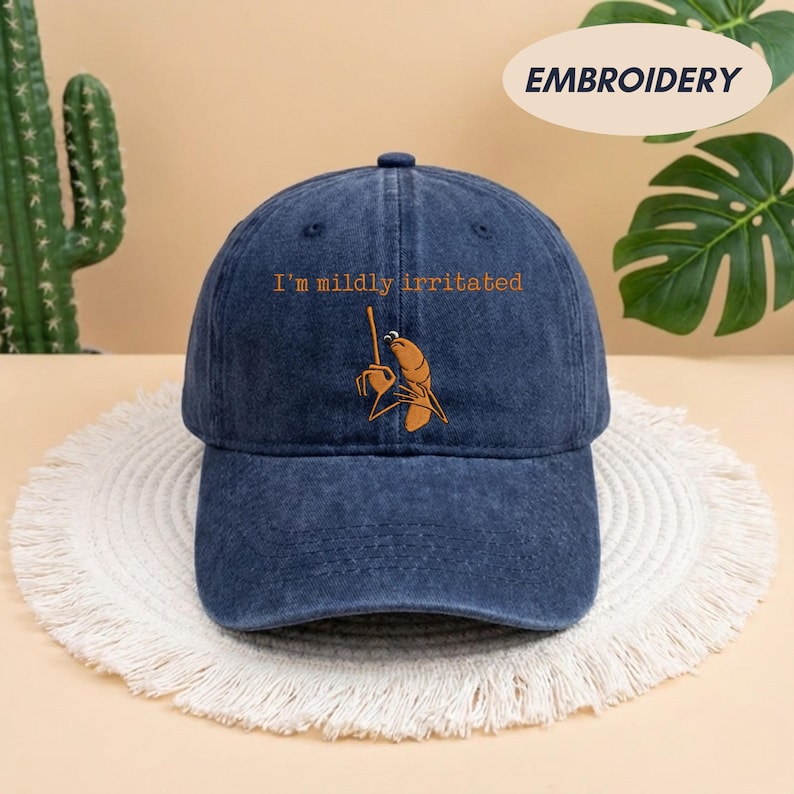 May include: A navy blue baseball cap with the embroidered phrase "I'm mildly irritated" and a cartoon image of a shrimp holding a tiny stick. The cap is sitting on a white, fringed mat.