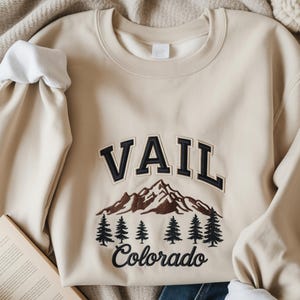Op de afbeelding: Een beige sweatshirt met ronde hals en het woord "VAIL" in zwarte letters boven een berg- en dennenboomontwerp. Het woord "Colorado" is eronder geborduurd. De mouwen hebben witte manchetten.