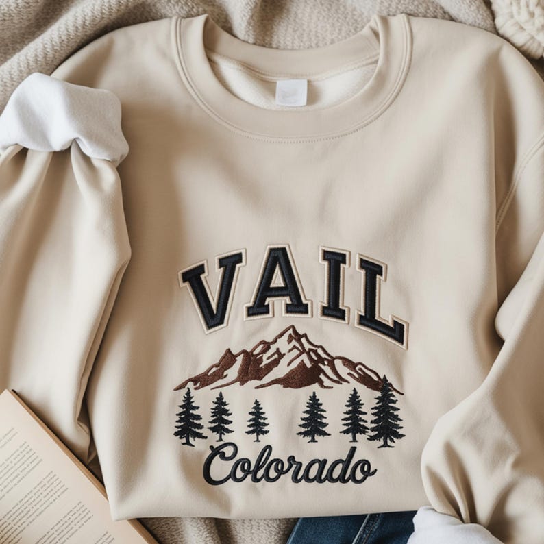 Op de afbeelding: Een beige sweatshirt met ronde hals en het woord "VAIL" in zwarte letters boven een berg- en dennenboomontwerp. Het woord "Colorado" is eronder geborduurd. De mouwen hebben witte manchetten.