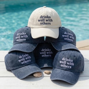 May include: A stack of baseball caps in various shades of blue and one cream-colored cap. Each cap has the embroidered text "drinks well with others" in a contrasting color. The caps are arranged in a pyramid shape.