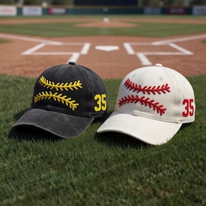 May include: Two baseball caps on green grass. One is black with yellow stitching and the number 35. The other is white with red stitching and the number 35. The caps are in front of a baseball field.