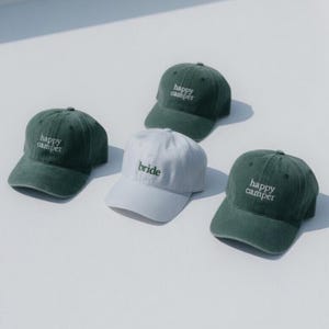May include: Four baseball caps are displayed on a white surface. Three are dark green with the words "happy camper" embroidered in white. The fourth cap is light blue with the word "bride" embroidered in white.