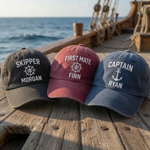 May include: Three baseball caps in charcoal, maroon, and navy blue. The charcoal cap features the text "SKIPPER MORGAN" with a ship wheel. The maroon cap displays "FIRST MATE FINN" with a ship wheel. The navy cap shows "CAPTAIN RYAN" with an anchor.