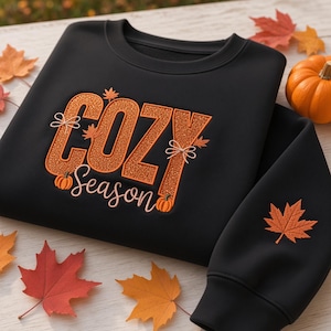 May include: A black crewneck sweatshirt featuring the orange glitter text COZY SEASON on the chest. The design includes small orange maple leaves, two thin white bows, and two small orange pumpkins. An orange maple leaf is embroidered on the sleeve.