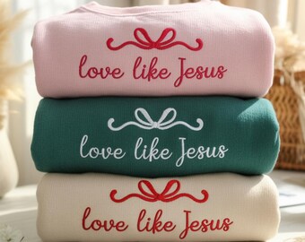 May include: Three stacked sweatshirts in pink, green, and beige. Each sweatshirt has the embroidered text "love like Jesus" with a bow design above the text. The sweatshirts are folded and stacked on top of each other.