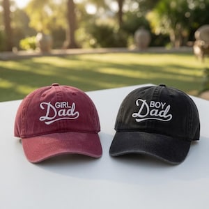 May include: Two baseball caps, one maroon and one black, sit side-by-side. The maroon cap has the text "Girl Dad" in white script, and the black cap has "Boy Dad" in white script. Both caps are on a white surface.